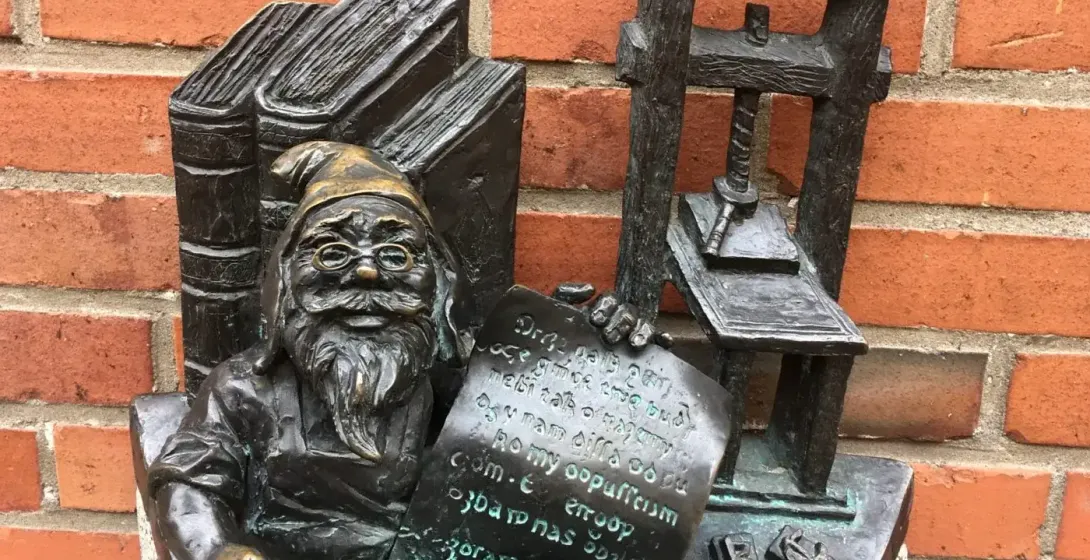 Brązowa statuetka krasnala w okularach, trzymającego dokument, obok książek i prasy drukarskiej, na tle czerwonej ceglanej...