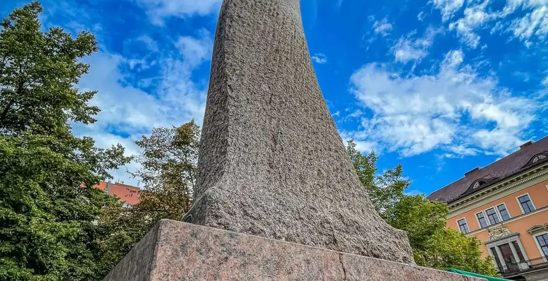 Wysoki, spiralny abstrakcyjny pomnik kamienny o chropowatej fakturze, na granitowej podstawie, pod błękitnym niebem z chmu...