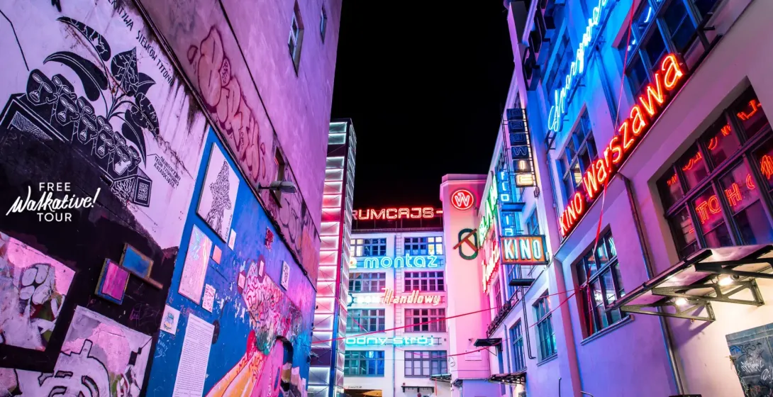 Wąska uliczka nocą z kolorowymi neonami na budynkach i ścianą pokrytą graffiti i muralami. Zaparkowane rowery.