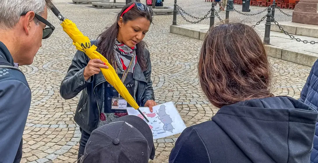 Przewodniczka z żółtym parasolem i mapą objaśnia trójce turystów na brukowanym placu. W tle kolorowe zabytkowe kamienice.