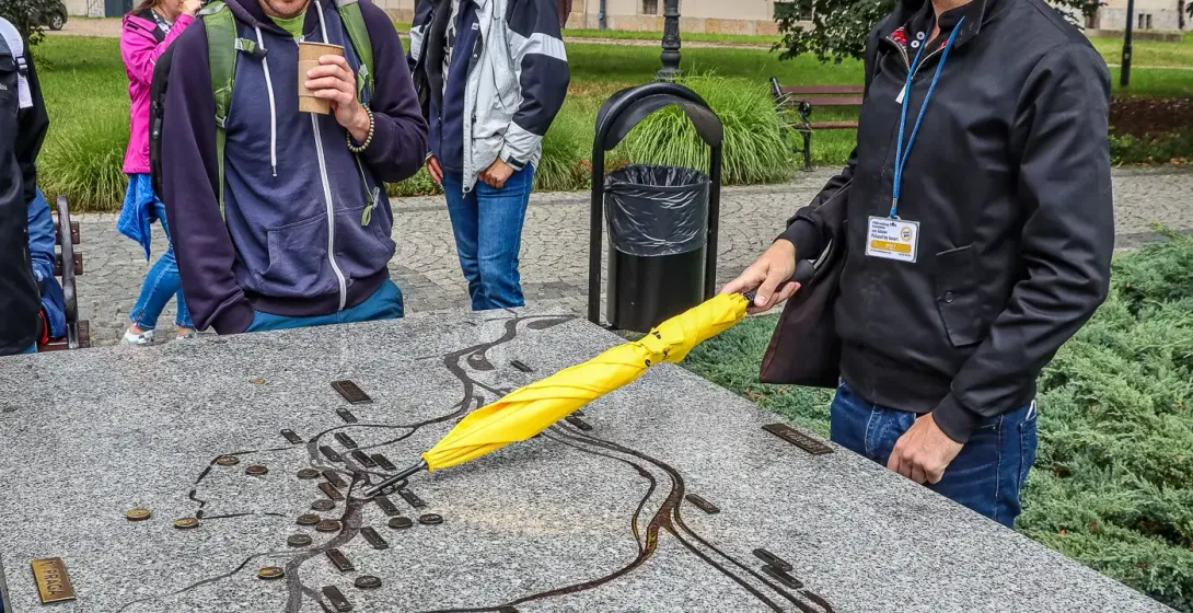 Tour guide with a yellow umbrella pointing at a tactile map of Wrocław while a small group of tourists listens.