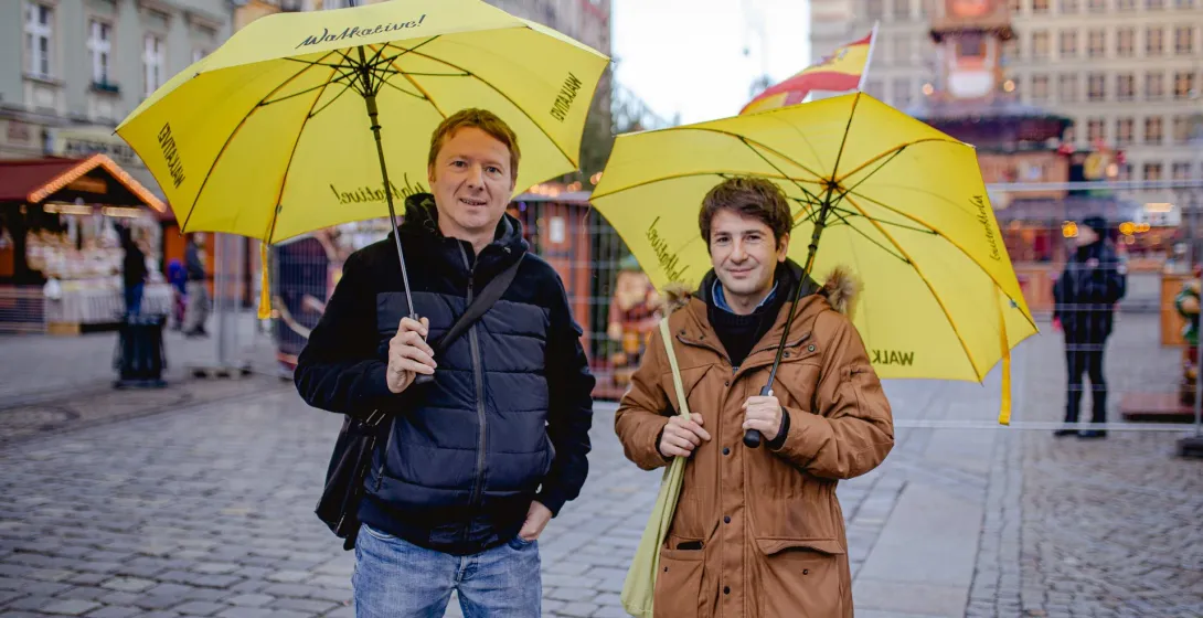 Dwóch mężczyzn z żółtymi parasolami Walkative! stoi na brukowanym placu z niewyraźnymi budynkami i straganami w tle.