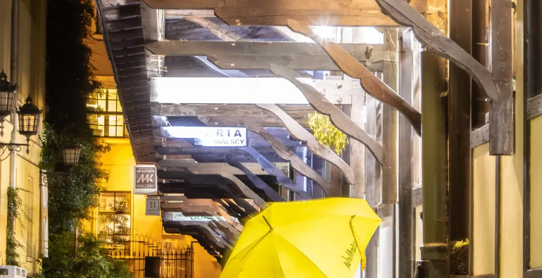 Żółty parasol Walkative! na brukowanej ulicy nocą, pod drewnianym pasażem oświetlonym jasnym światłem.
