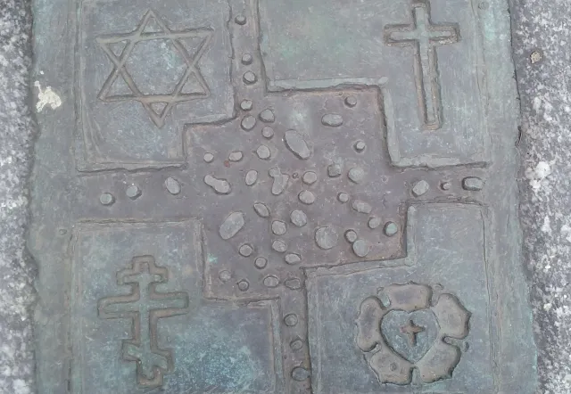 Bronze plaque depicting four religious symbols: Star of David, Christian cross, Orthodox cross, and a heart with a cross.