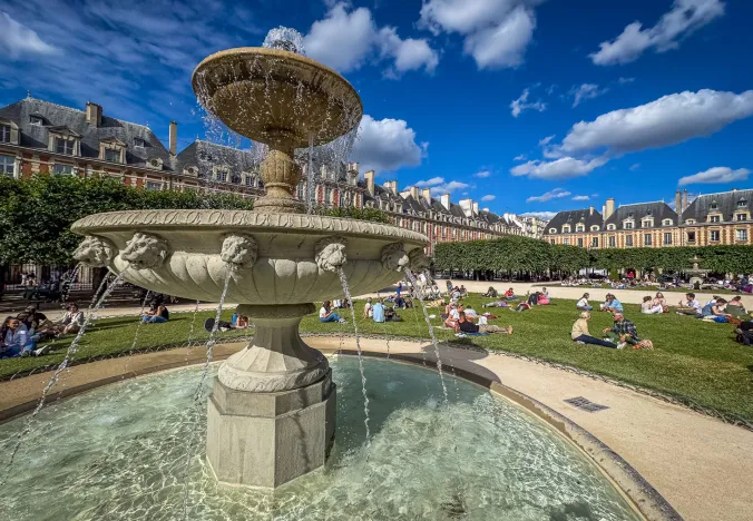 Ein malerischer Brunnen auf dem Place des Vosges im Marais, Paris, umgeben von Menschen, die einen sonnigen Tag genießen.