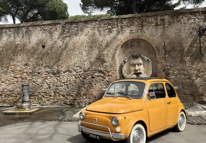 Żółty Fiat 500 zaparkowany w pobliżu rzymskiej ściany z unikalną fontanną.