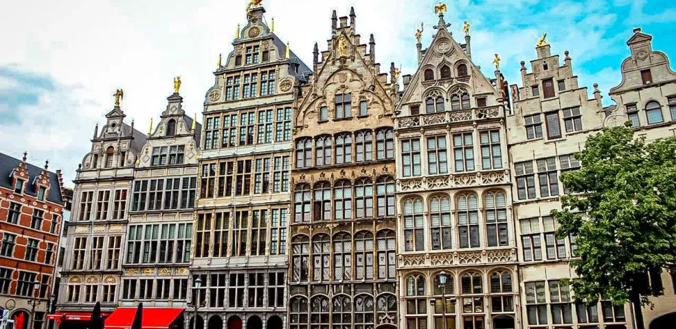 Historische Zunfthäuser mit Treppengiebeln und goldenen Statuen säumen einen Kopfsteinpflasterplatz in Antwerpen. Passanten g