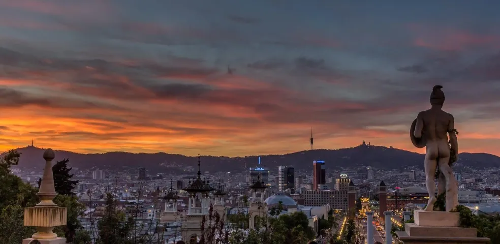 View of Barcelona city skyline at sunset with a statue in the foreground and Tibidabo mountain in the background.