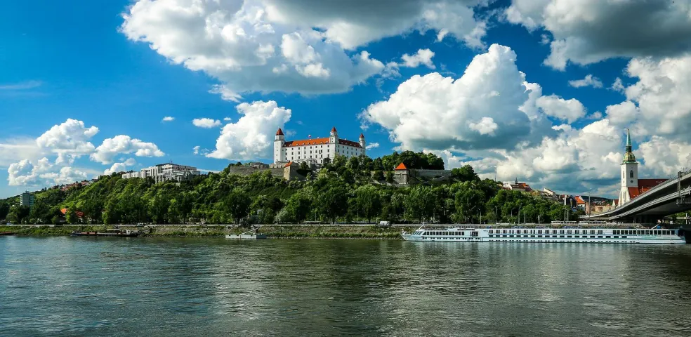 Castillo de Bratislava en una colina verde sobre el río Danubio, con un crucero, puente y aguja de la Catedral de San Martín.