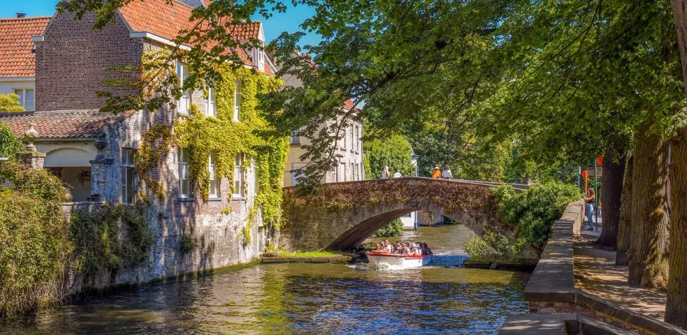 Łódź turystyczna z pasażerami na kanale w Brugii, przepływająca pod kamiennym mostem obok budynku pokrytego bluszczem.