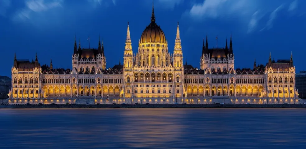 El Parlamento húngaro iluminado de noche, visto a través del oscuro río Danubio bajo un cielo nublado.