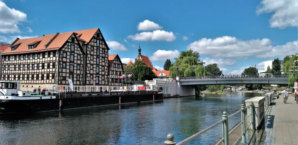 Half-timbered buildings with red roofs on the riverbank, a long barge, a concrete bridge, and a pedestrian path under a blue