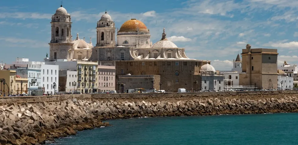 View of Cádiz Cathedral's domes and bell towers alongside other city buildings from across the turquoise sea.