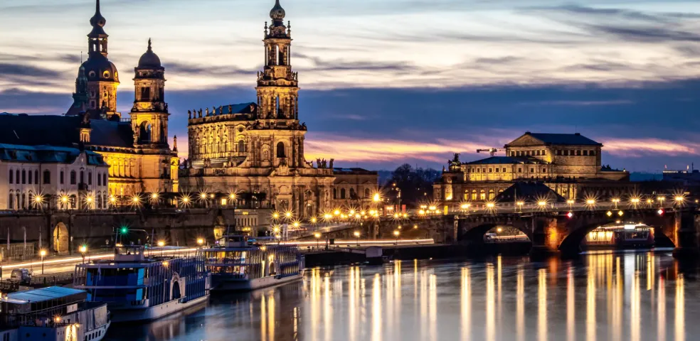 Dresden skyline at twilight over the Elbe River, with illuminated historic buildings and reflections on the water.