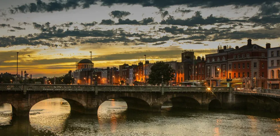 Dublin cityscape at sunset with Ha'penny Bridge over the River Liffey, glowing warm light, and historical buildings.