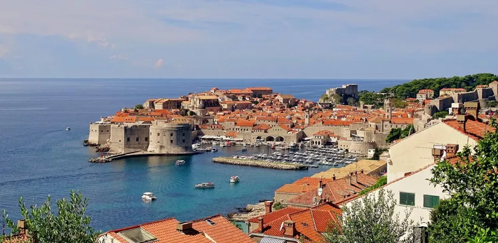 Elevated view of Dubrovnik's Old Town, featuring red-tiled roofs, medieval city walls, a harbor with boats, and the Adriatic