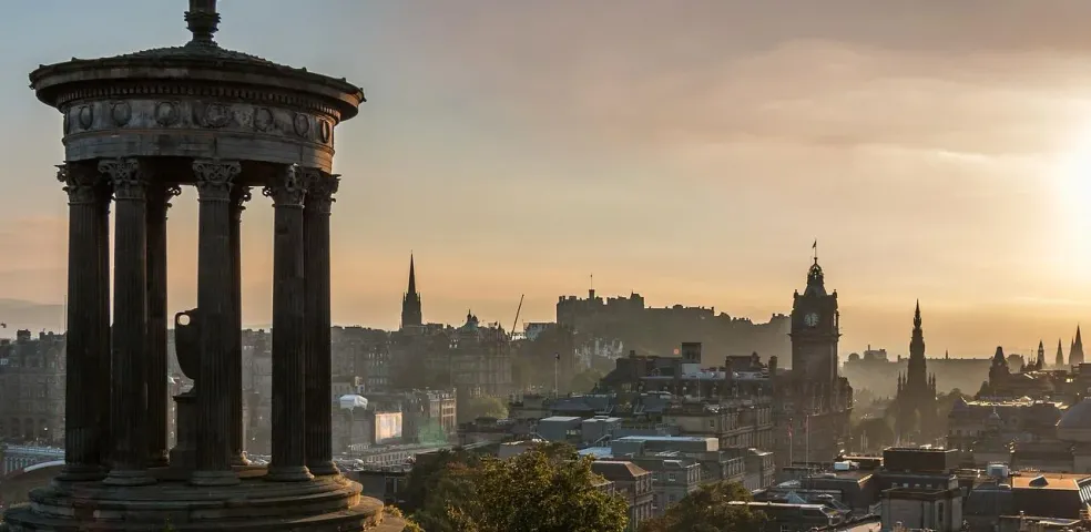 Dugald Stewart Monument foreground, Edinburgh Castle, and city skyline at sunrise with hazy sky.