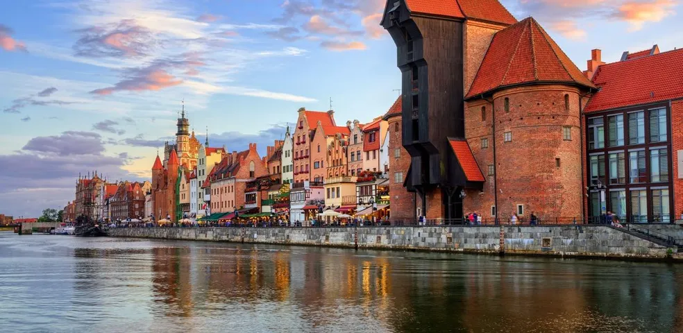Historic waterfront buildings and a large wooden crane reflecting in the Motława River at sunset in Gdańsk.