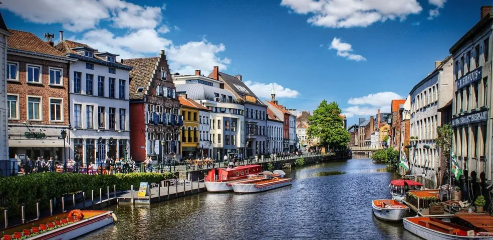 A sunny view of a canal in Ghent lined with historic, colorful buildings and several tour boats docked along the side.