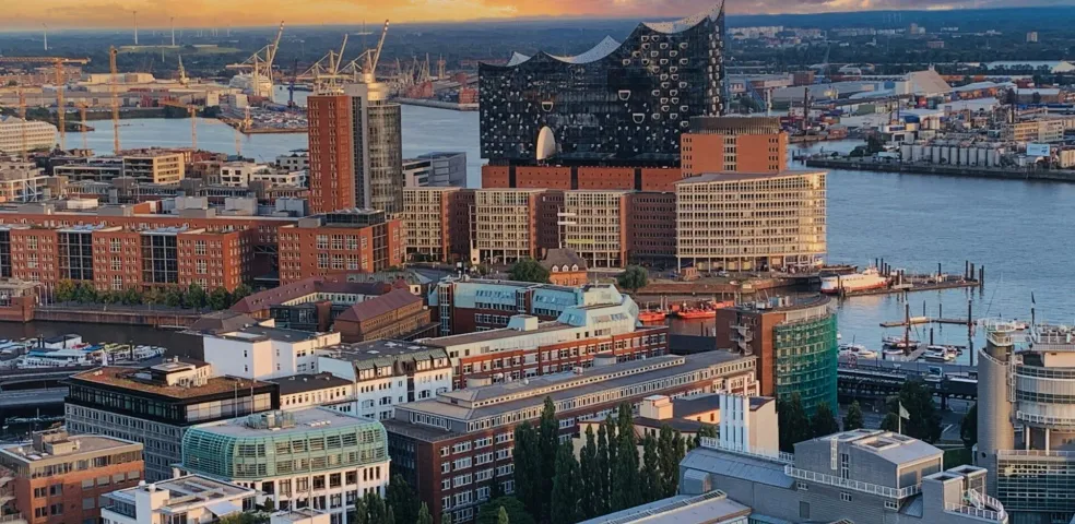 Luftaufnahme des Hamburger Hafens bei Sonnenuntergang mit der Elbphilharmonie, Backsteingebäuden und Hafenkranen am Wasser.