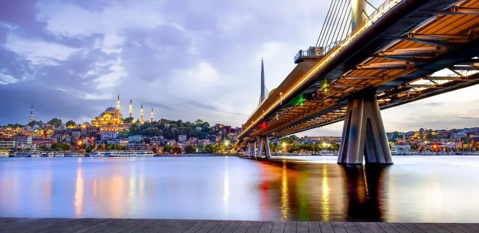 Illuminated Golden Horn Metro Bridge over water at dusk, with the Süleymaniye Mosque and Istanbul cityscape in the background
