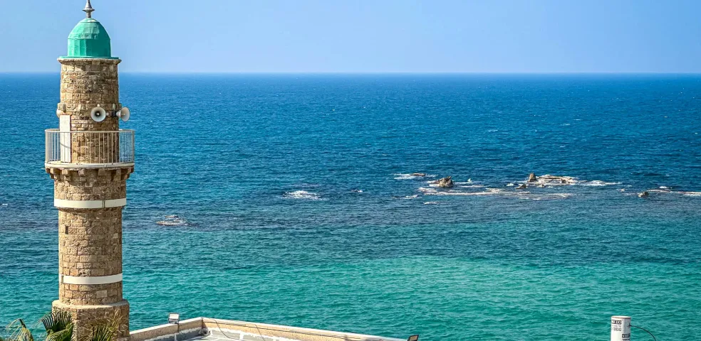 Minarete de piedra con cúpula verde y balcón, dominando el mar Mediterráneo turquesa, con tejados y follaje verde.