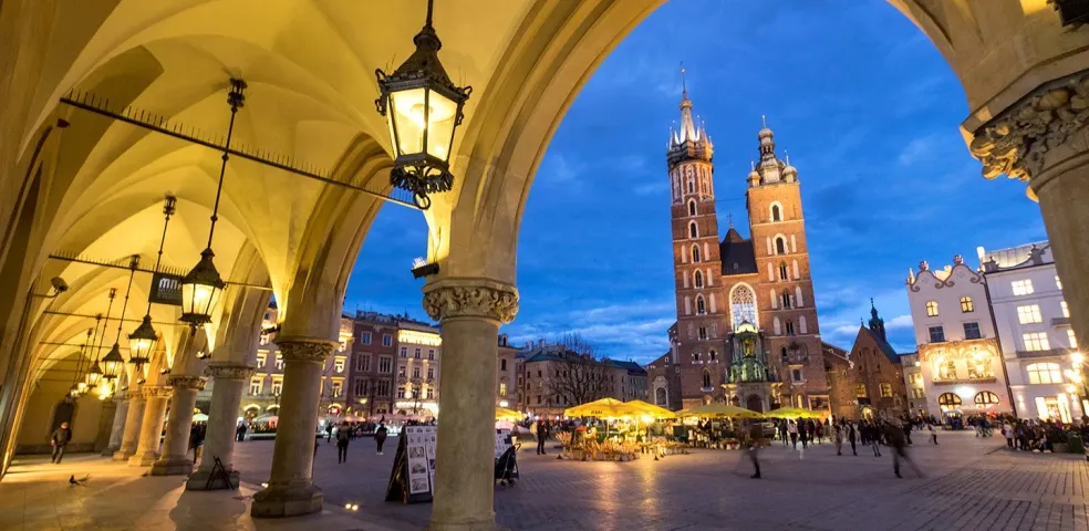 View from an illuminated archway of the Krakow Main Market Square with St. Mary's Basilica at dusk.