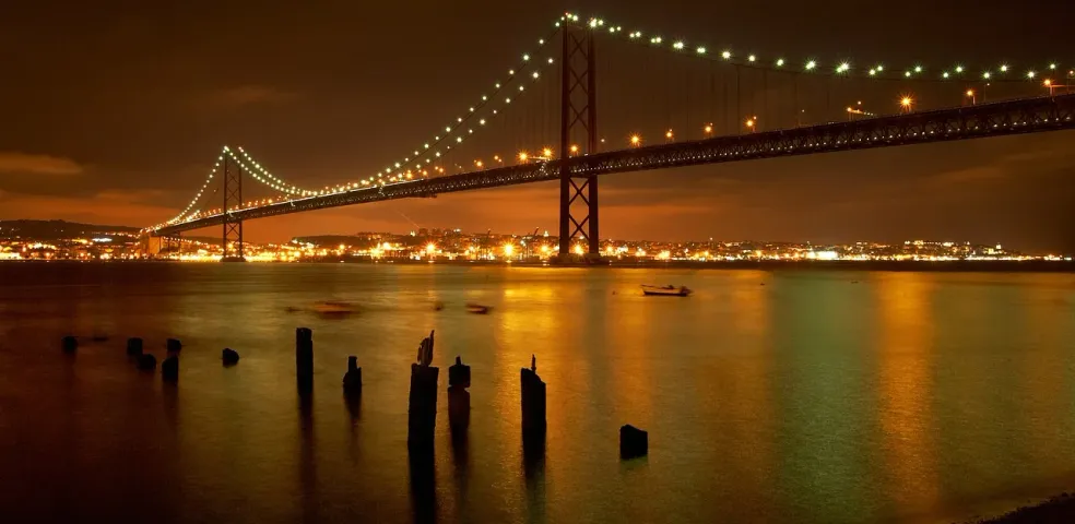 25 de Abril Bridge illuminated at night over the Tagus River, with city lights and reflections on the water.