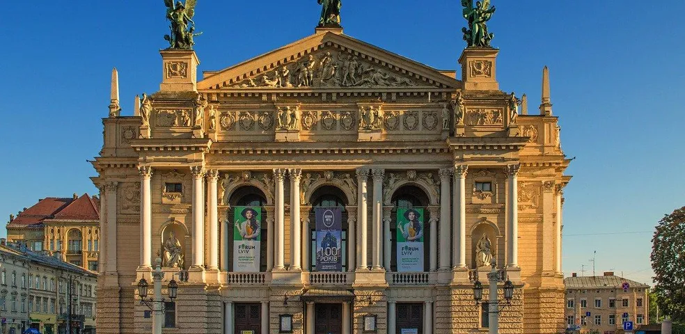 Fassade der Lwiwer Oper mit Säulen, Skulpturen auf dem Dach und Bannern. Klarer blauer Himmel. Menschen gehen im Vordergrund.