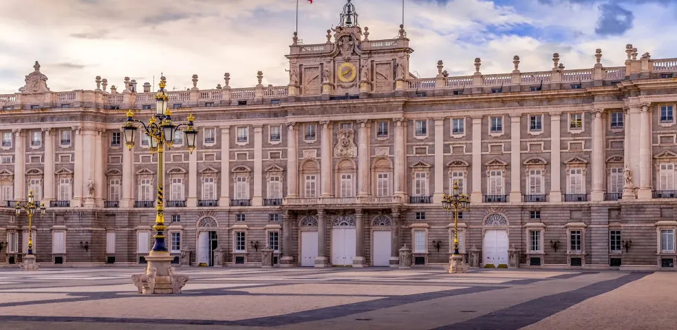 Exterior view of the Royal Palace of Madrid under a dramatic sky, with cobblestone plaza and lampposts.