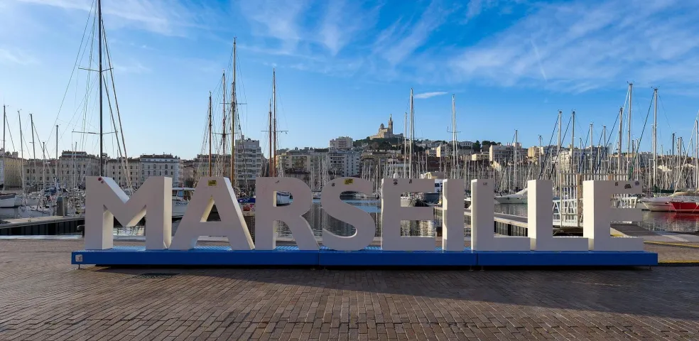Große MARSEILLE-Buchstaben auf blauem Sockel am Vieux-Port, mit Segelbooten und Gebäuden. Notre-Dame de la Garde auf dem Hüge