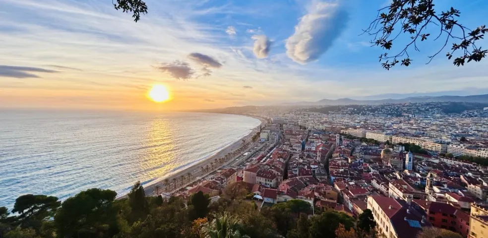 Panoramic view of Nice, France at sunset, showing the curved coastline, city buildings, and a bright sun over the sea.