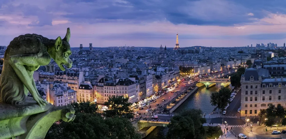 Gárgola de piedra en un edificio con vistas al horizonte de París al anochecer, con la Torre Eiffel iluminada y luces de la c