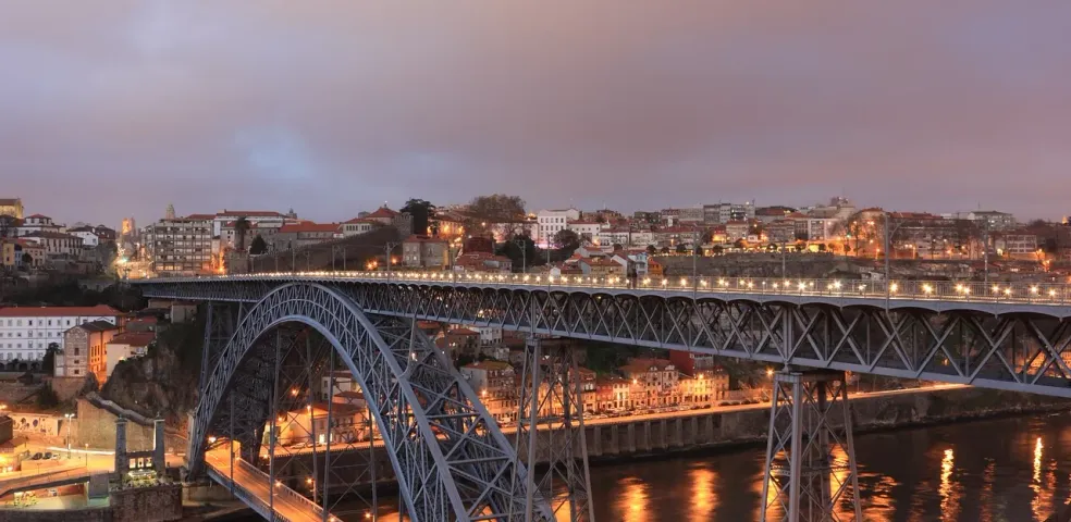 Large metal arch bridge with two decks illuminated at dusk over a river, with city buildings on both sides.