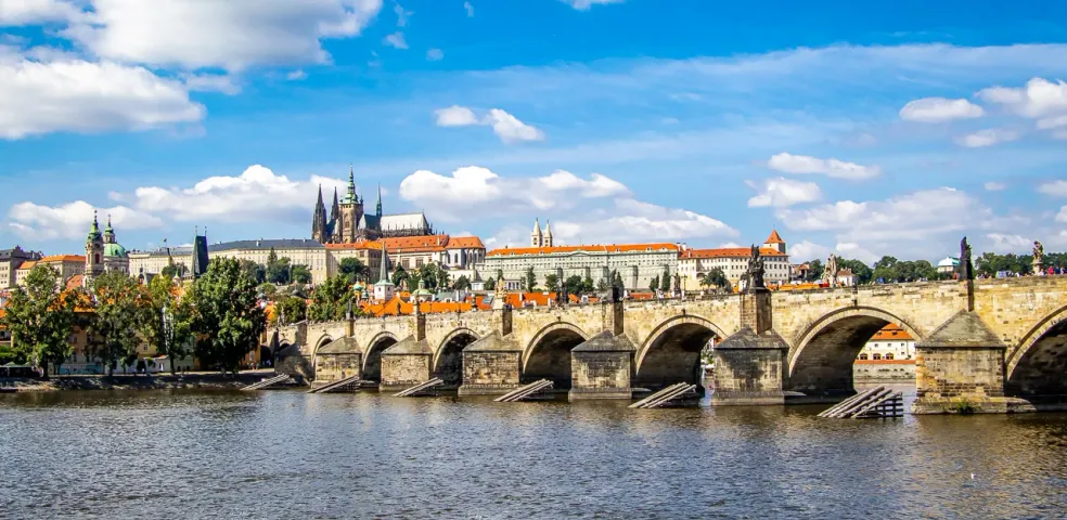Blick auf die Karlsbrücke mit Statuen und Bögen über einem Fluss, Prager Burg am fernen linken Ufer unter blauem Himmel mit W