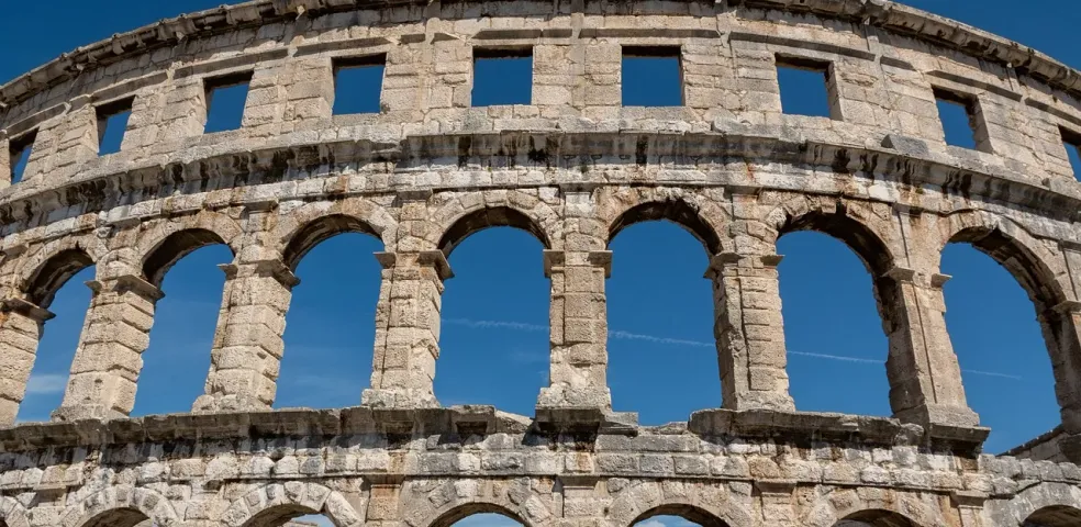 Exterior view of the Pula Arena, an ancient Roman amphitheater with multiple arched levels and windows against a clear blue s