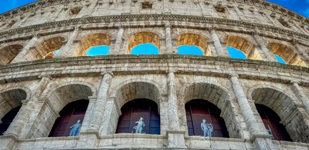 Vista exterior del Coliseo en Roma, mostrando múltiples niveles de arcos de piedra, columnas y estatuas en los arcos inferior
