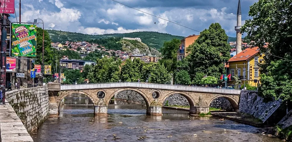 Historische Steinbogenbrücke über einen Fluss in Sarajevo, mit einem Minarett, Gebäuden und grünen Hügeln unter bewölktem Him