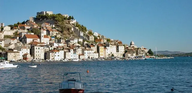 Sibenik old town and hilltop fortress from the sea, showing historic buildings with red roofs, boats, and a clear blue sky.