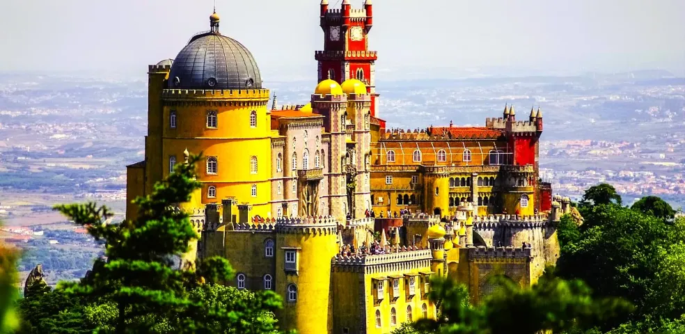 Colorful Pena Palace in Sintra, Portugal, featuring yellow, red, and grey sections with domes and towers, seen from afar thro
