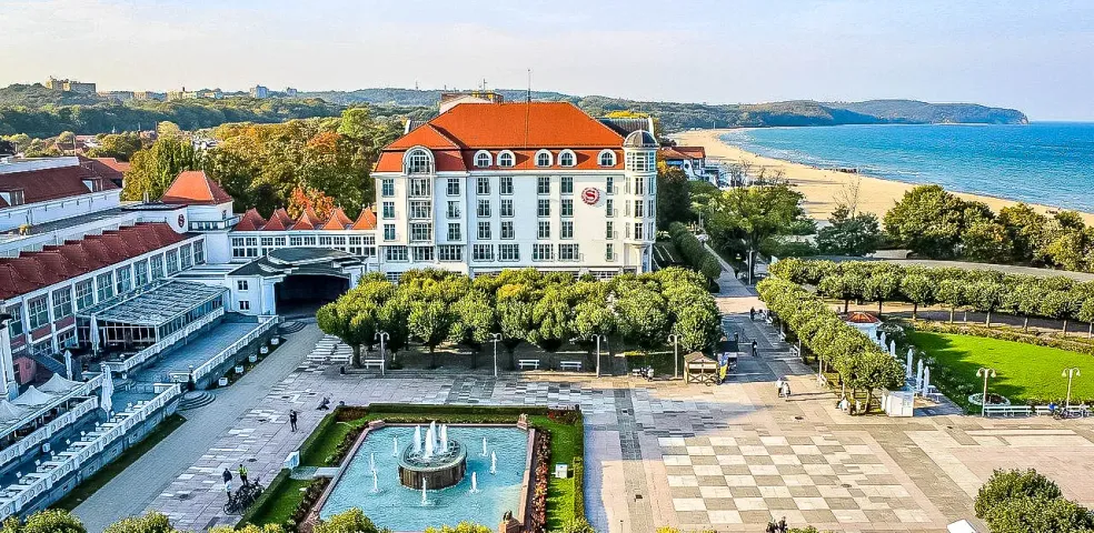 Aerial view of Grand Hotel Sopot with a square, fountain, green trees, sandy beach, and Baltic Sea under a blue sky.