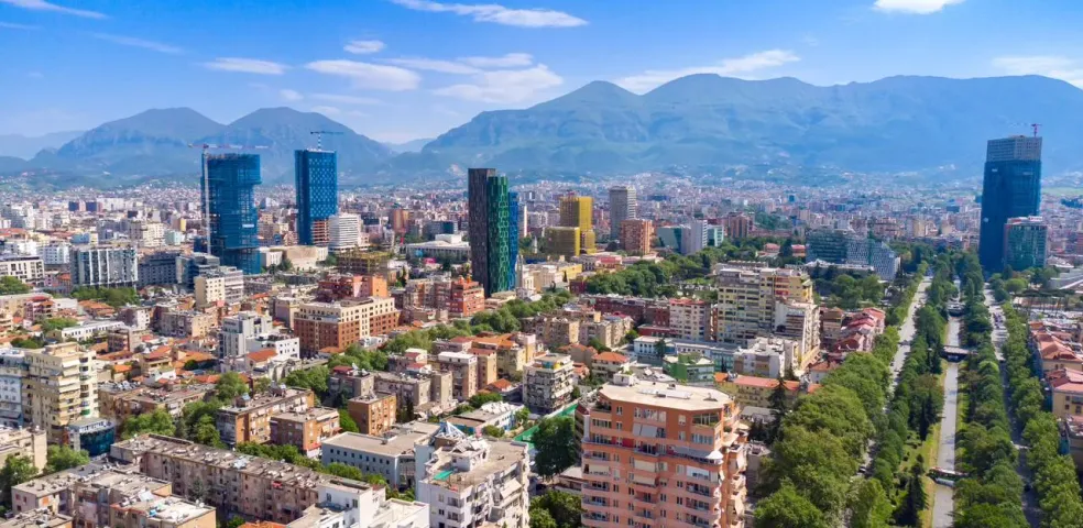 Aerial view of Tirana city with modern high-rise buildings, residential areas, a tree-lined boulevard, and mountains.