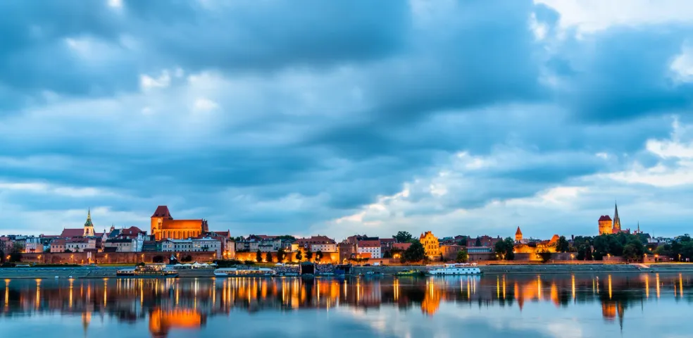 Panorama Torunia o zmierzchu, z historycznymi budynkami podświetlonymi wzdłuż rzeki, odbijającymi się w spokojnej wodzie pod