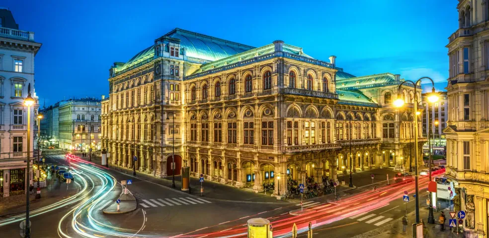 Vienna State Opera at twilight with light trails from cars on the street below and a crescent moon.