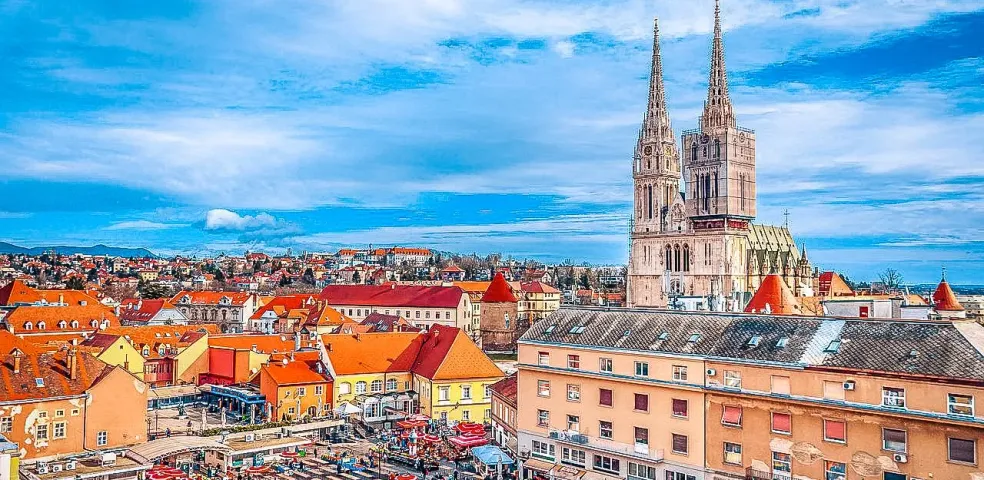 Aerial view of Zagreb with the Cathedral featuring two spires, partially scaffolded, and a bustling outdoor market with red u