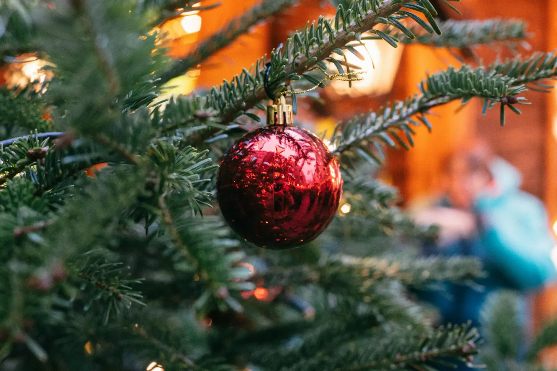 Rote Weihnachtskugel an einem Tannenzweig vor verschwommenen warmen Lichtern im Hintergrund.