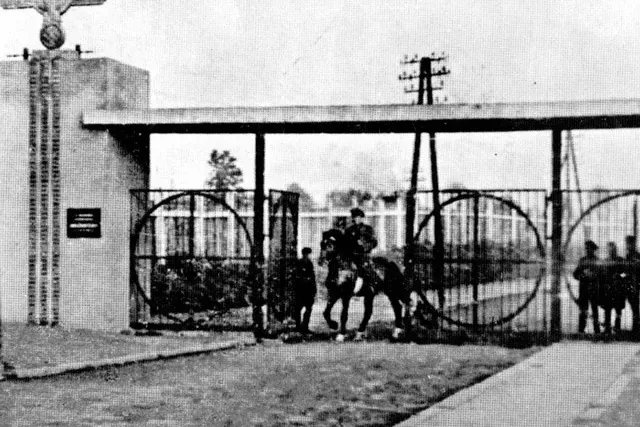 Black and white photo of a camp entrance gate with a Nazi eagle emblem, men in uniform, and circular metalwork.