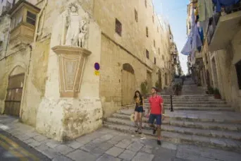 Dos personas bajan escalones de piedra en una calle estrecha de La Valeta, con edificios antiguos y una estatua en la esqu...