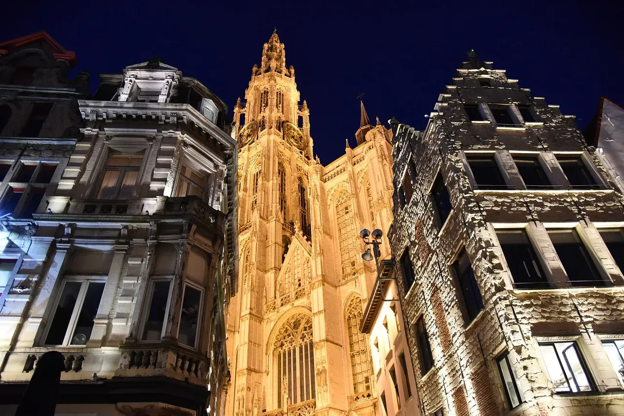 Beleuchteter gotischer Kathedralenturm bei Nacht, umgeben von historischen europäischen Gebäuden unter dunklem Himmel.