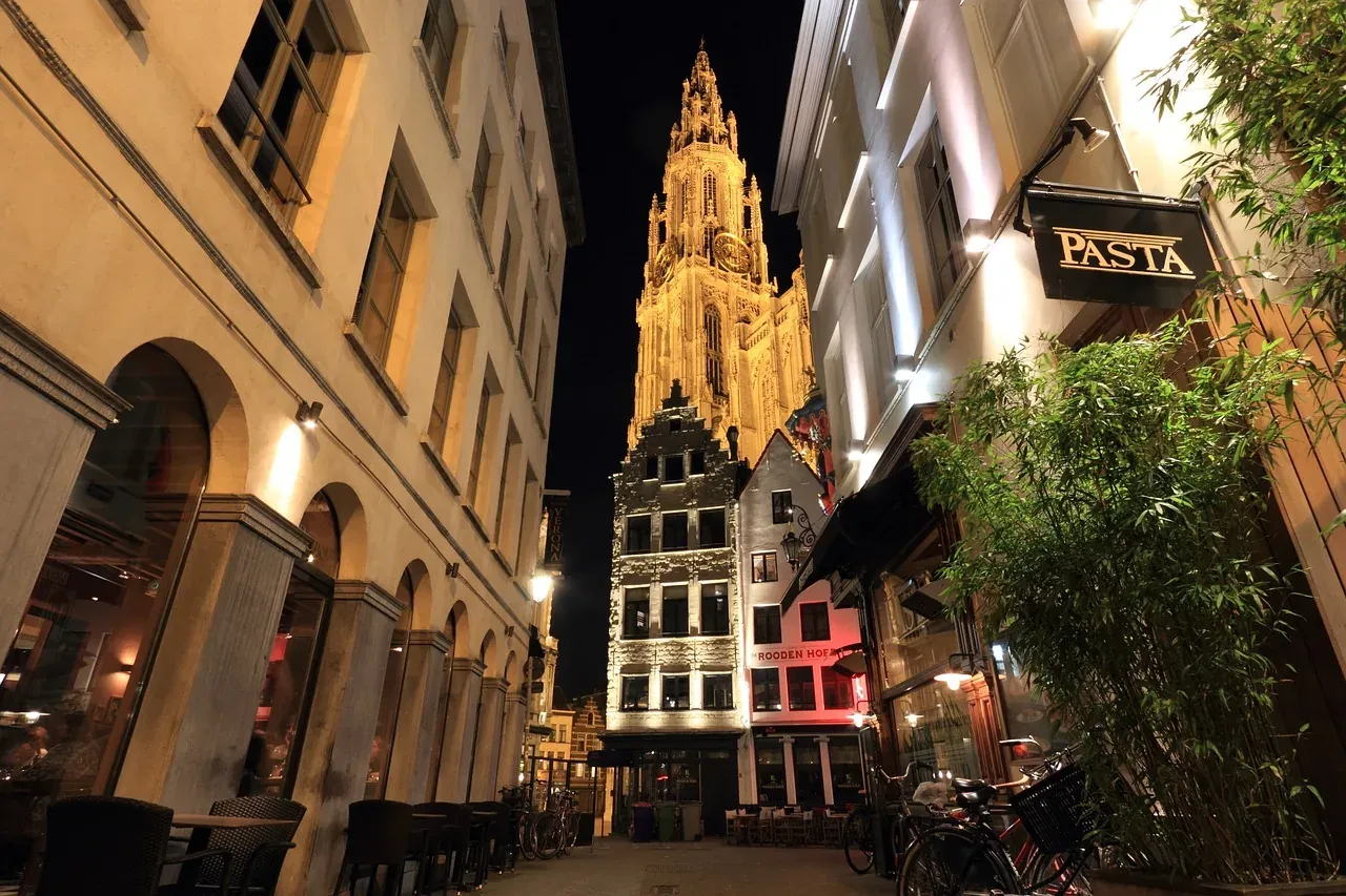 Der beleuchtete Turm der Liebfrauenkathedrale Antwerpen bei Nacht, gesehen von einer engen Gasse mit Gebäuden.